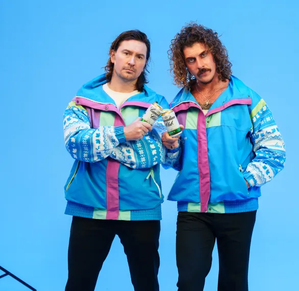 Two people wearing vibrant, retro ski jackets pose against a blue background. holding a can, with a spotlight visible in the corner, conveying a playful 80s vibe.