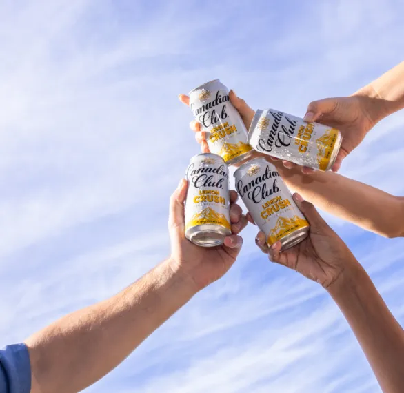 Four hands clink cans of Canadian Club Lemon Crush against a bright blue sky, celebrating togetherness and relaxation.