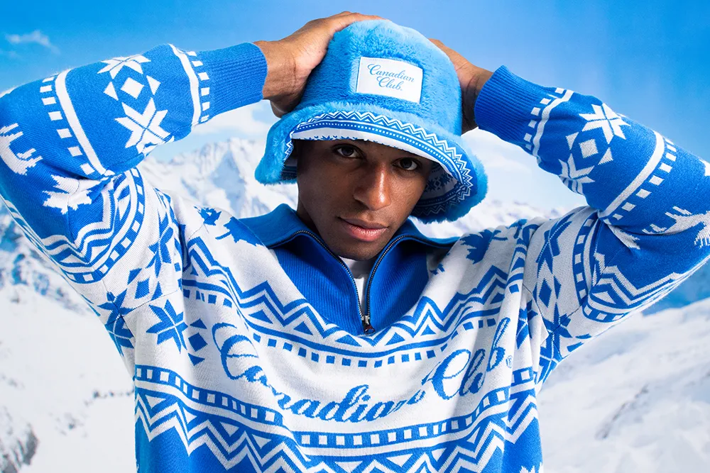 A person wearing a blue and white patterned sweater and matching hat stands against a snowy mountain backdrop.