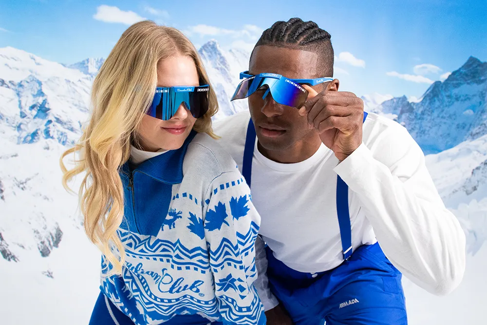 A man and a woman pose playfully in front of a snowy mountain backdrop, dressed in stylish winter attire.