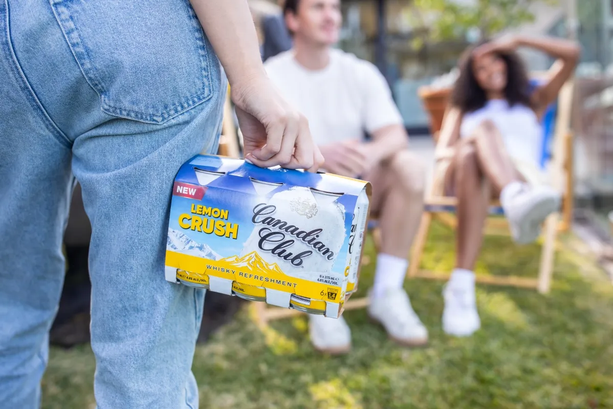A person in jeans holds a pack of Canadian Club Lemon Crush drinks, with two friends relaxing in the background on lawn chairs.