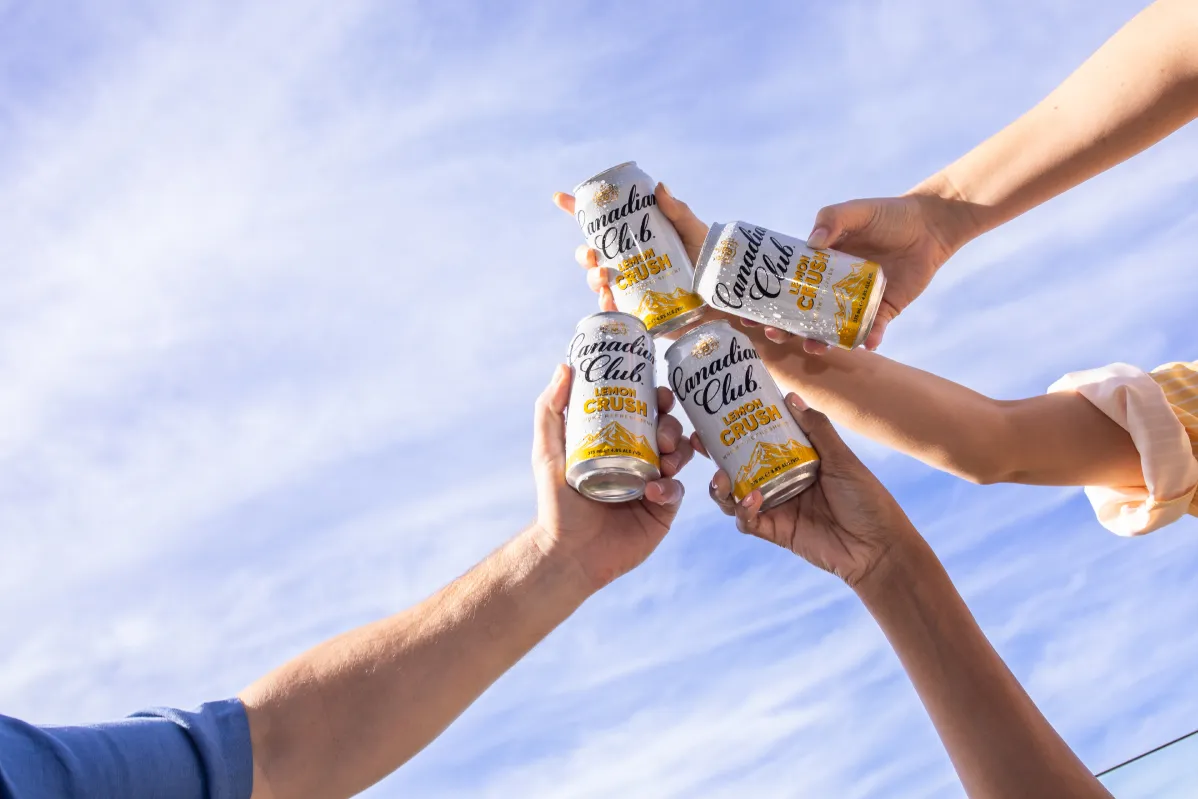 Four hands clink cans of Canadian Club Lemon Crush against a bright blue sky, celebrating togetherness and relaxation.