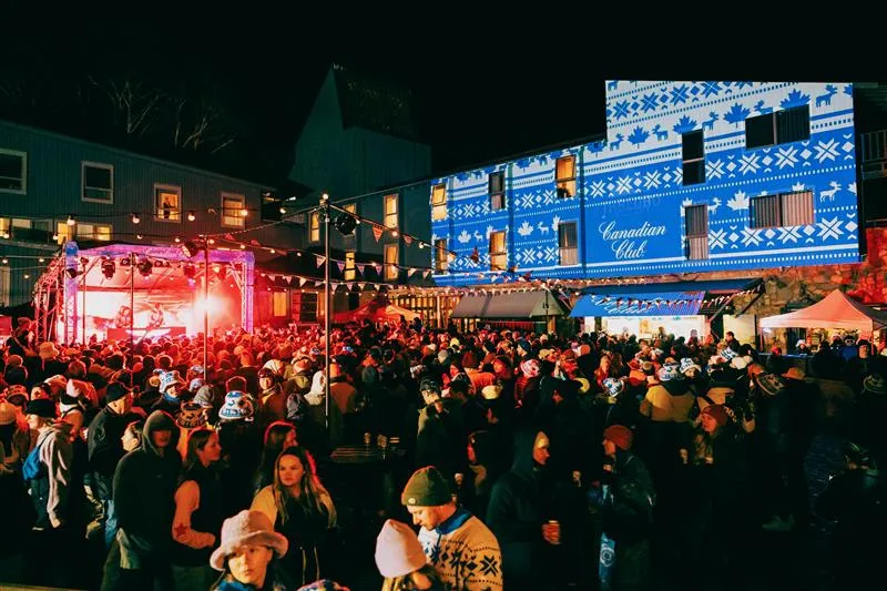 Large crowd attending an outdoor nighttime event with a lit stage and festive lights; a building in the background is decorated with blue, white, and maple leaf patterns.