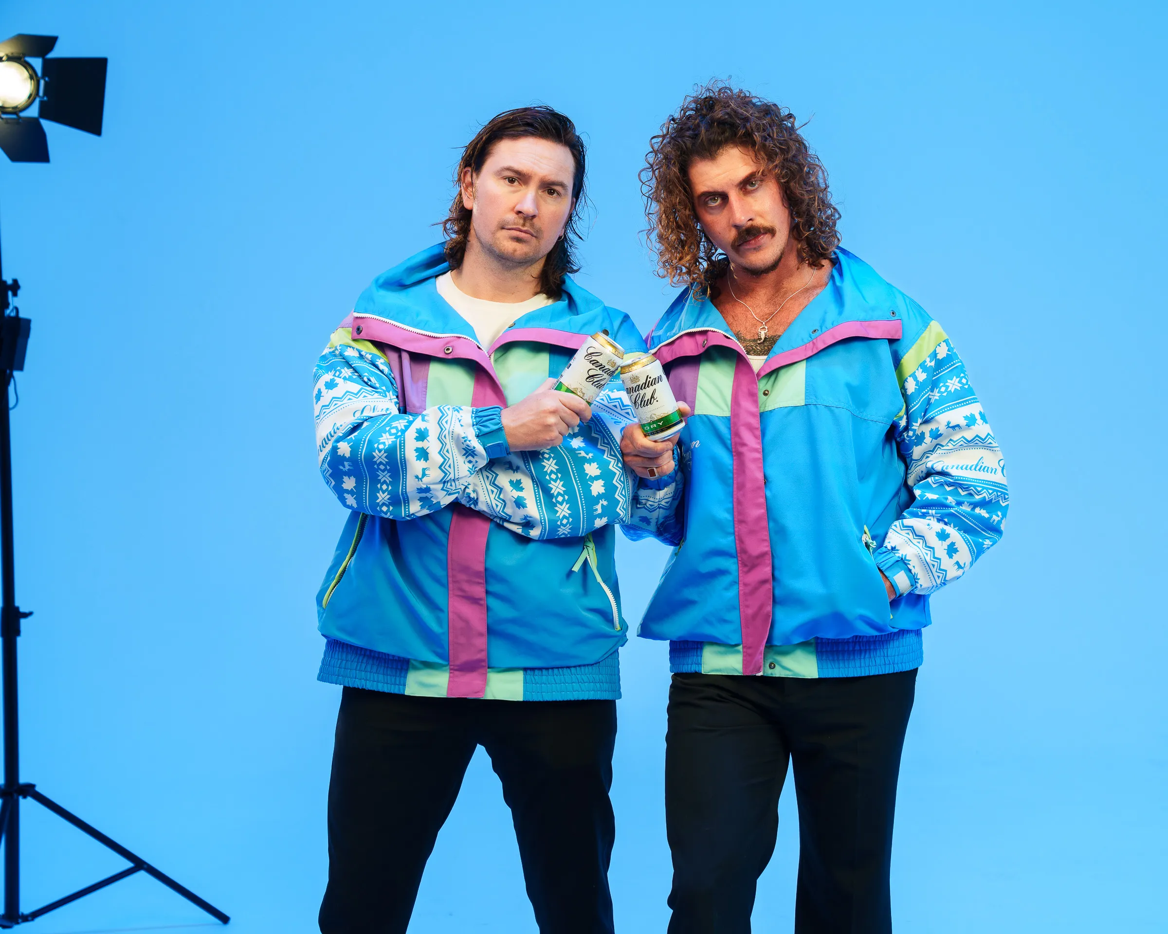 Two people wearing vibrant, retro ski jackets pose against a blue background. holding a can, with a spotlight visible in the corner, conveying a playful 80s vibe.